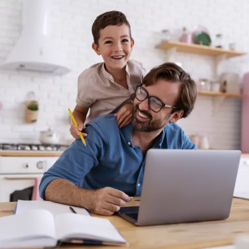 A joyful scene with a smiling man working on his laptop in the kitchen of his house, while his young son laughs and playfully jumps on his back, creating a heartwarming and family-oriented home workspace