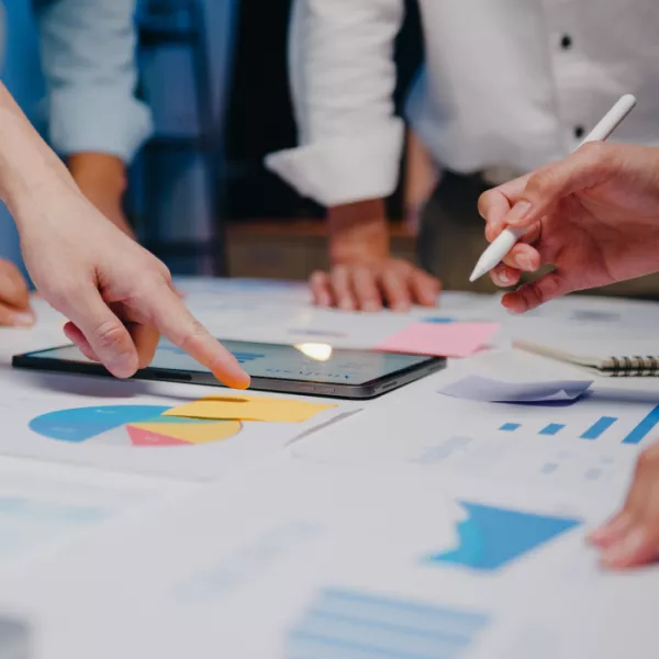 A close-up shot of the hands of several people standing around a table, engaged in a discussion about strategies and charts, highlighting a collaborative and analytical meeting