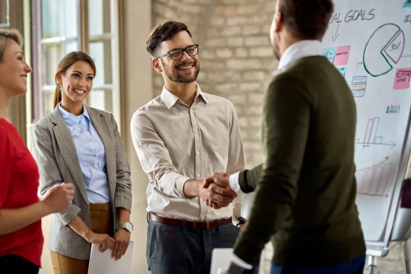 A group of professionals, wearing smiles with two individuals engaged in a handshake, while a background board displays charts, conveying a positive and successful business interaction