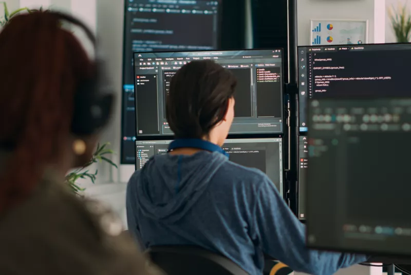 A person seen from behind, seated at a computer with multiple screens, engrossed in coding or programming work, portraying a focused and productive workspace