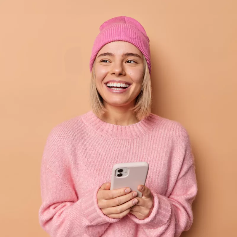 A young woman dressed in pink, holding a cellphone in her hands against a peach background, exuding joy and a vibrant, youthful vibe